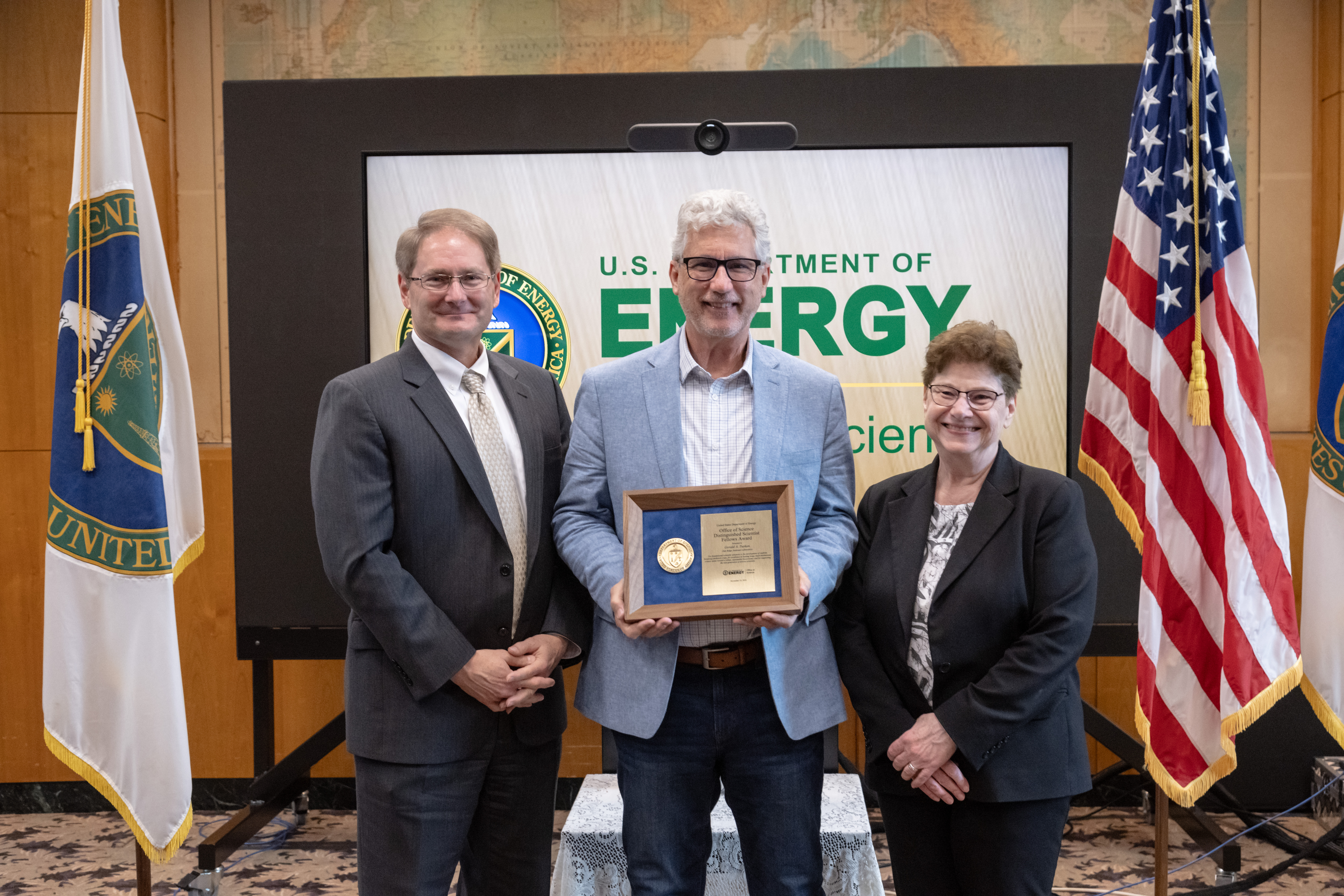 Three well-dressed, smiling people stand in front of a presentation board that says "U.S. Department of Energy" The person in the center holds an award plaque.