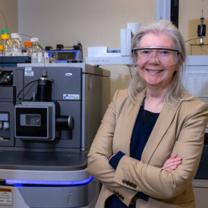 Researcher wearing saftey glasses poses with a mass spectrometer in a research lab.