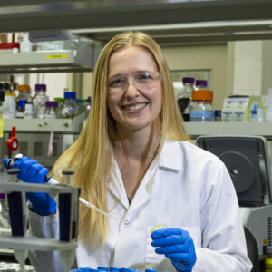 Scientist in a lab coat and gloves using a pipette in a laboratory setting.