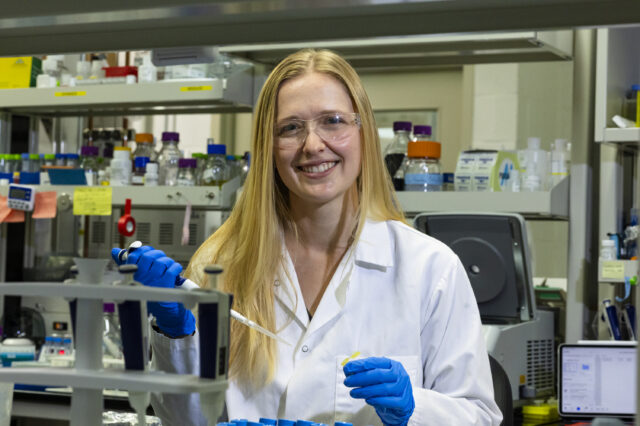 Scientist in a lab coat and gloves using a pipette in a laboratory setting.