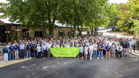 A large group of people is gathered outdoors, standing closely together for a group photo. They are holding a bright green banner with the logo and name "CBI Center for Bioenergy Innovation" printed on it. The background includes a building with trees and greenery, suggesting the photo was taken at an outdoor event or conference. The group appears diverse in terms of age, gender, and attire.