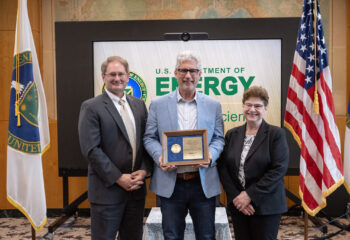 Three well-dressed, smiling people stand in front of a presentation board that says "U.S. Department of Energy" The person in the center holds an award plaque.