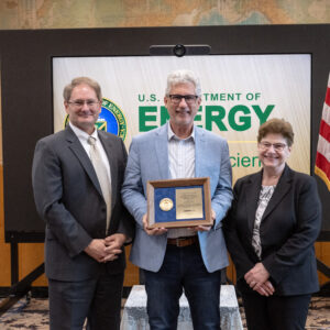 Three well-dressed, smiling people stand in front of a presentation board that says "U.S. Department of Energy" The person in the center holds an award plaque.