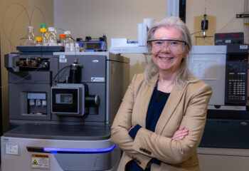Researcher wearing saftey glasses poses with a mass spectrometer in a research lab.