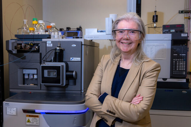 Researcher wearing saftey glasses poses with a mass spectrometer in a research lab.